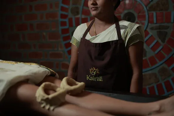 Therapist in a Kairali apron beside a patient with herbal dough rings on both knees