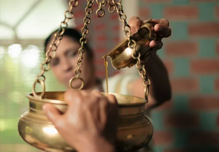 Therapist pouring herbal oil into a brass dhara vessel suspended on chains